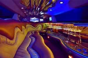 Luxurious NJ limousine interior with curved leather seats, blue and gold lighting, a ceiling mirror, multiple TVs, and a bar lined with empty champagne glasses—ideal for premier limo service Newark experiences.