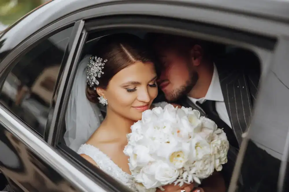 A bride holding a bouquet of white flowers smiles while sitting in a sleek black car service Newark, as her groom in a suit gently leans in behind her.