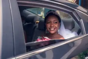 A bride wearing a tiara and veil smiles while sitting in the backseat of a black car service Newark, holding a bouquet of flowers and looking out the open window.