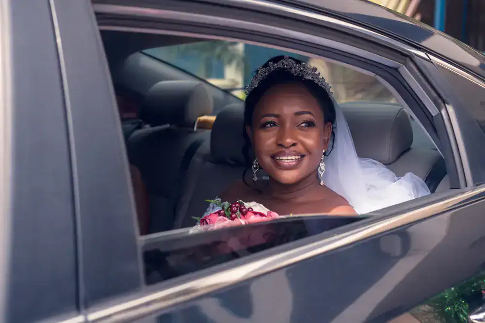A bride wearing a tiara and veil smiles while sitting in the backseat of a black car service Newark, holding a bouquet of flowers and looking out the open window.