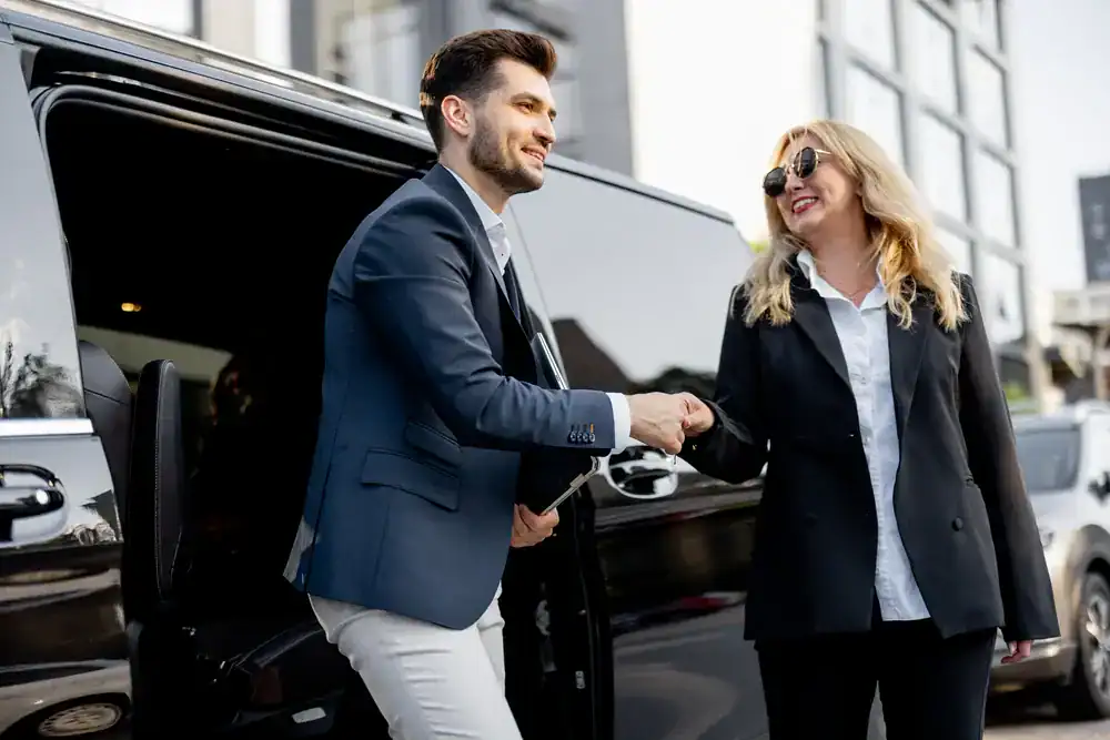 A man in a suit helps a smiling woman in business attire out of a sleek black car, shaking her hand. Both appear professional, representing a premier black car service Newark, and are standing outside near modern buildings.