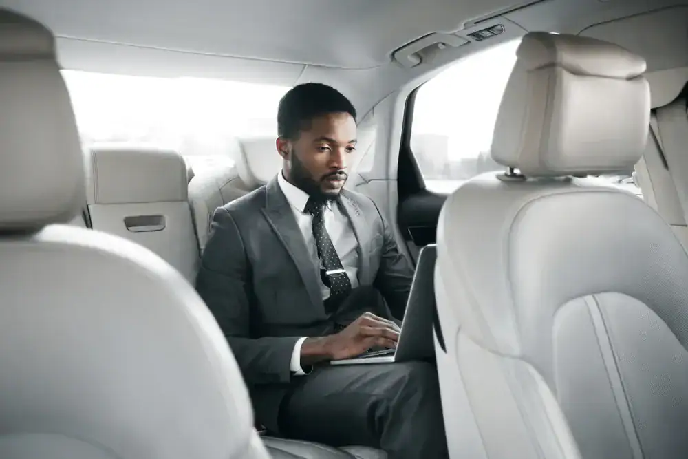 A man in a suit sits in the backseat of a modern, light-colored car, working intently on his laptop—enjoying the comfort and privacy of a black car service Newark for his business commute.