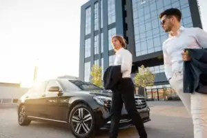Two men in business attire stand beside a black limo service Newark car, parked in front of a modern glass office building, with sunlight shining in the background.