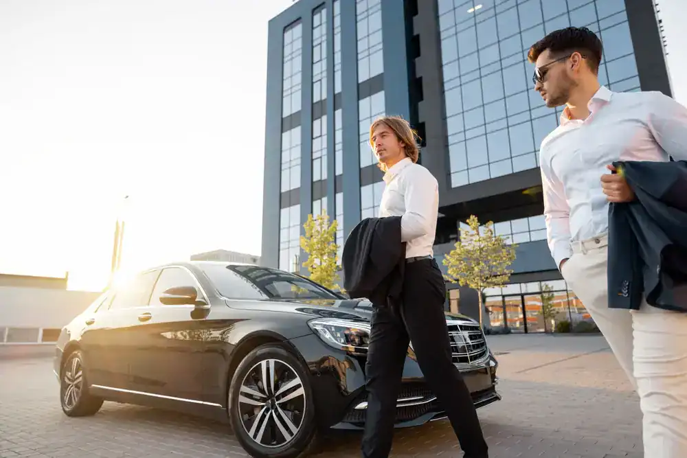 Two men in business attire stand beside a black limo service Newark car, parked in front of a modern glass office building, with sunlight shining in the background.
