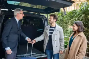 A man in a suit greets a smiling couple beside an open car trunk, holding their suitcase as they stand together—capturing the professional touch of a black car service Newark at the start or end of their journey.