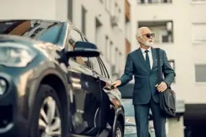 An older man in a suit and sunglasses stands by a black car, ready to enter. Carrying a shoulder bag outside a modern building, he appears prepared for his black car service Newark experience.
