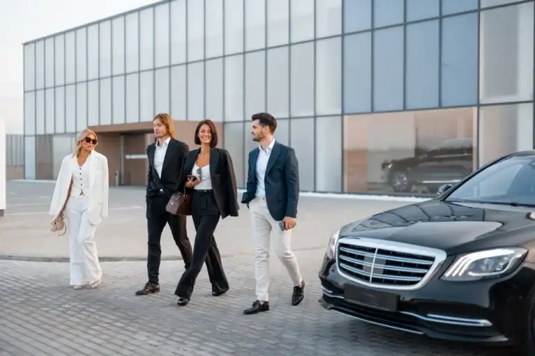 Four professionally dressed people walk together on a paved area near a modern glass building, smiling. A sleek black car, representing premium black car service Newark, waits in the foreground.