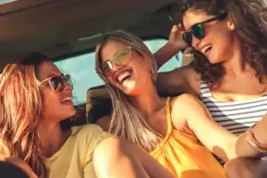 Three young women wearing sunglasses sit close together, smiling and laughing inside a black car service Newark, NJ, with the sun shining in, creating a joyful and carefree atmosphere.