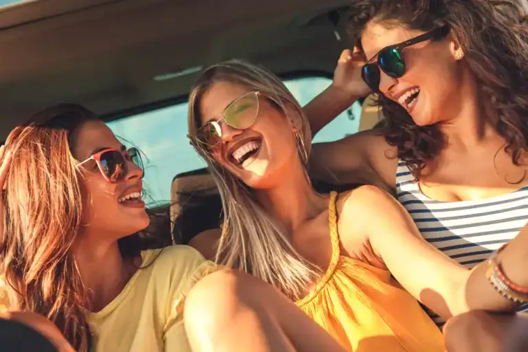 Three young women wearing sunglasses sit close together, smiling and laughing inside a black car service Newark, NJ, with the sun shining in, creating a joyful and carefree atmosphere.
