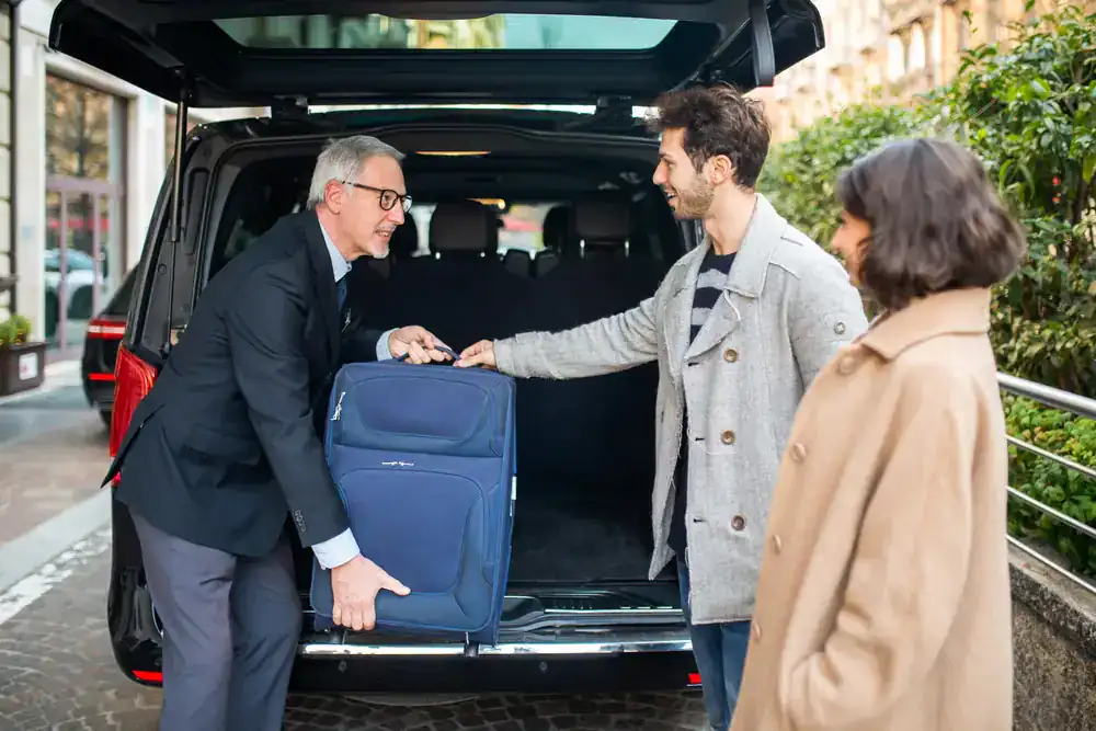 A man in a suit helps load a blue suitcase into the trunk of a black van from a black car service Newark while a casually dressed man and a woman in a tan coat stand nearby on a city street.