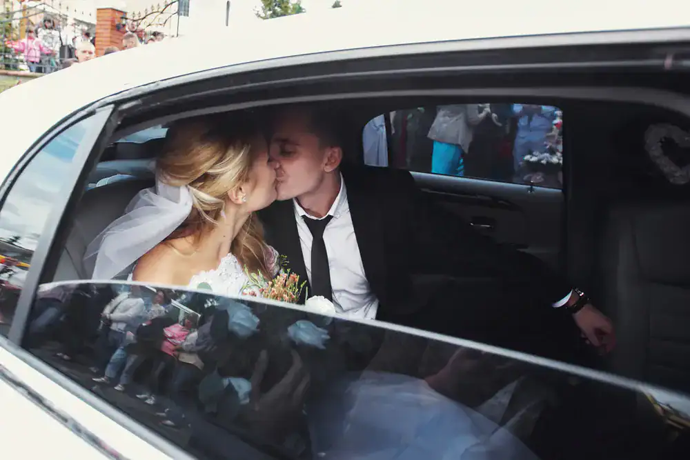 Bride and groom sit in the back seat of a black car service Newark, kissing, with wedding guests and onlookers reflected in the window outside.