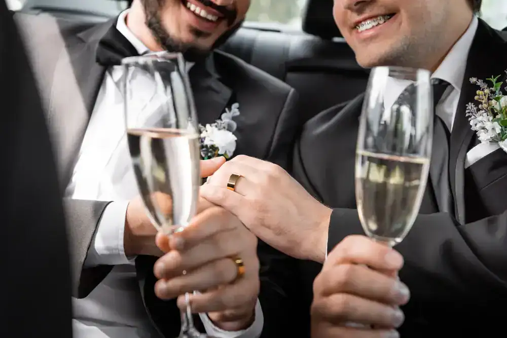 Two grooms in tuxedos smile and hold hands, both wearing wedding rings, while holding champagne glasses, celebrating together after arriving in style with a black car service Newark offers.