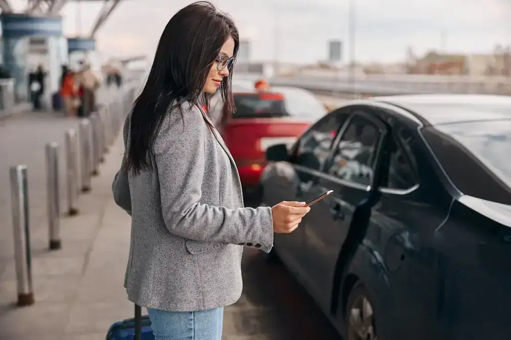 A woman with long dark hair, wearing glasses, a gray blazer, and jeans, stands by a row of cars at an outdoor curbside in NJ, looking at her smartphone and holding a suitcase, perhaps waiting for her black car service Newark to arrive.