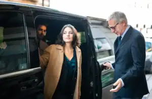 A woman in a tan coat steps out of a black car service Newark, escorted by a man in a suit and glasses. Another man is visible inside the vehicle. The urban setting suggests it could be outside a building in NJ.