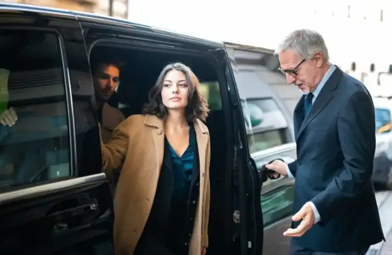 A woman in a tan coat steps out of a black car service Newark, escorted by a man in a suit and glasses. Another man is visible inside the vehicle. The urban setting suggests it could be outside a building in NJ.