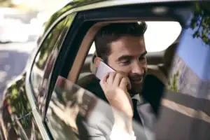 A man in a suit sits in the back seat of a car, smiling while talking on his smartphone. Sunlight filters through the window, reflecting some trees and the outdoors on the car's shiny surface.