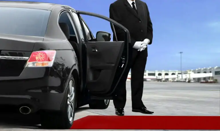 A chauffeur in a black suit and white gloves stands by an open car door next to a black luxury sedan, representing NJ's premier black car service Newark, with a red carpet rolled out and an airport terminal in the background.