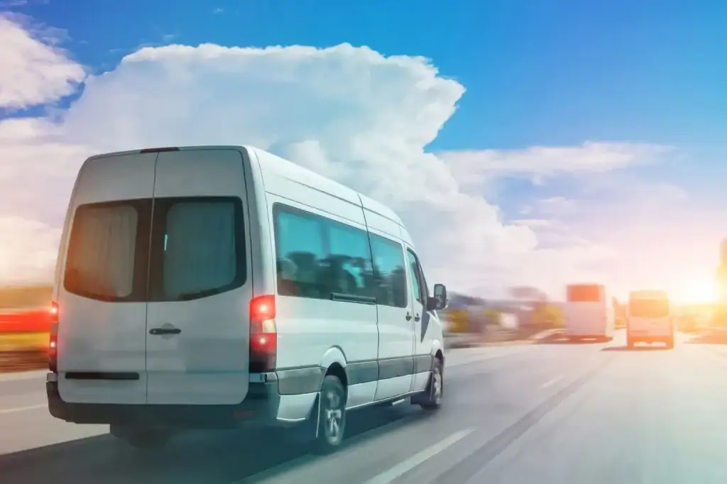 A white passenger van drives on a sunny highway with a few other vehicles, blue sky, and large clouds in the background.