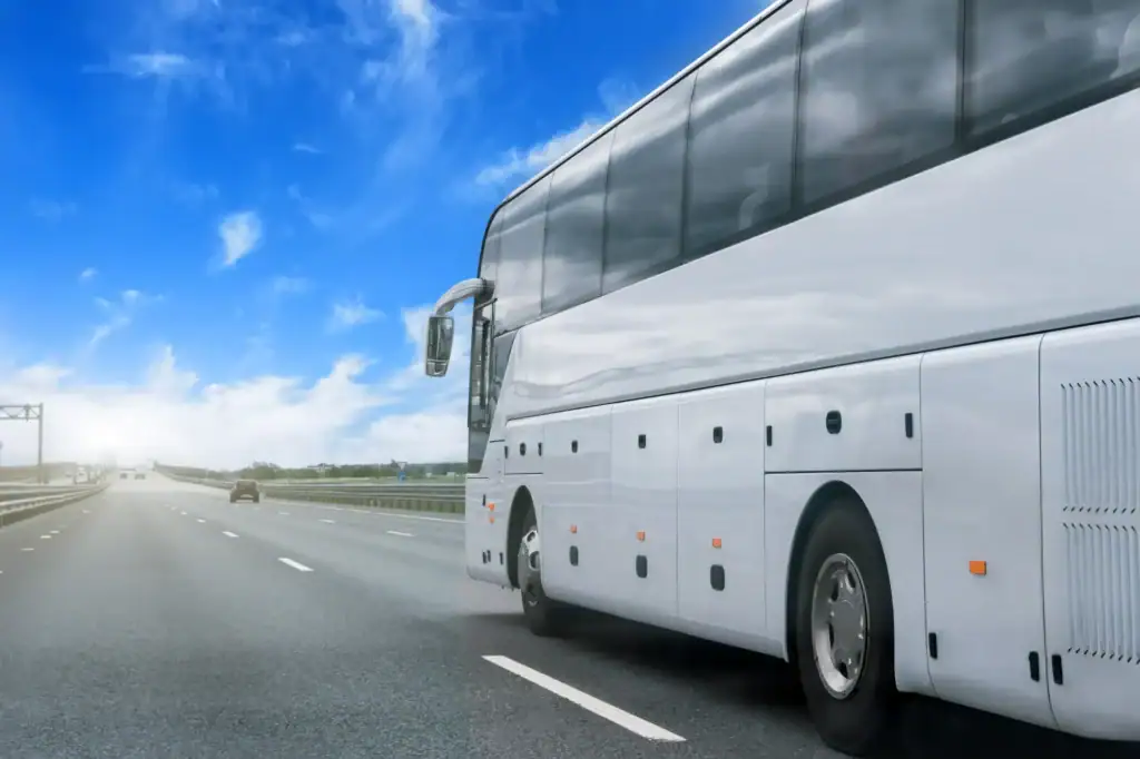 A large white coach bus drives down a nearly empty highway under a bright blue sky with scattered clouds, as a black car service from Newark, NJ cruises in the opposite lane.