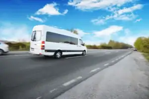 A white passenger van drives along a highway under a bright blue sky with scattered clouds, passing green trees, other vehicles, and a black car service Newark, NJ on a clear day.