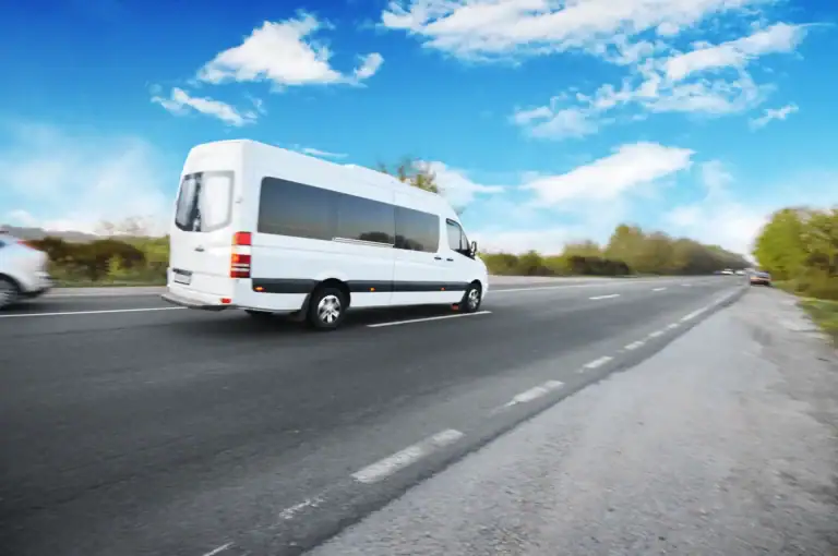 A white passenger van drives along a highway under a bright blue sky with scattered clouds, passing green trees, other vehicles, and a black car service Newark, NJ on a clear day.