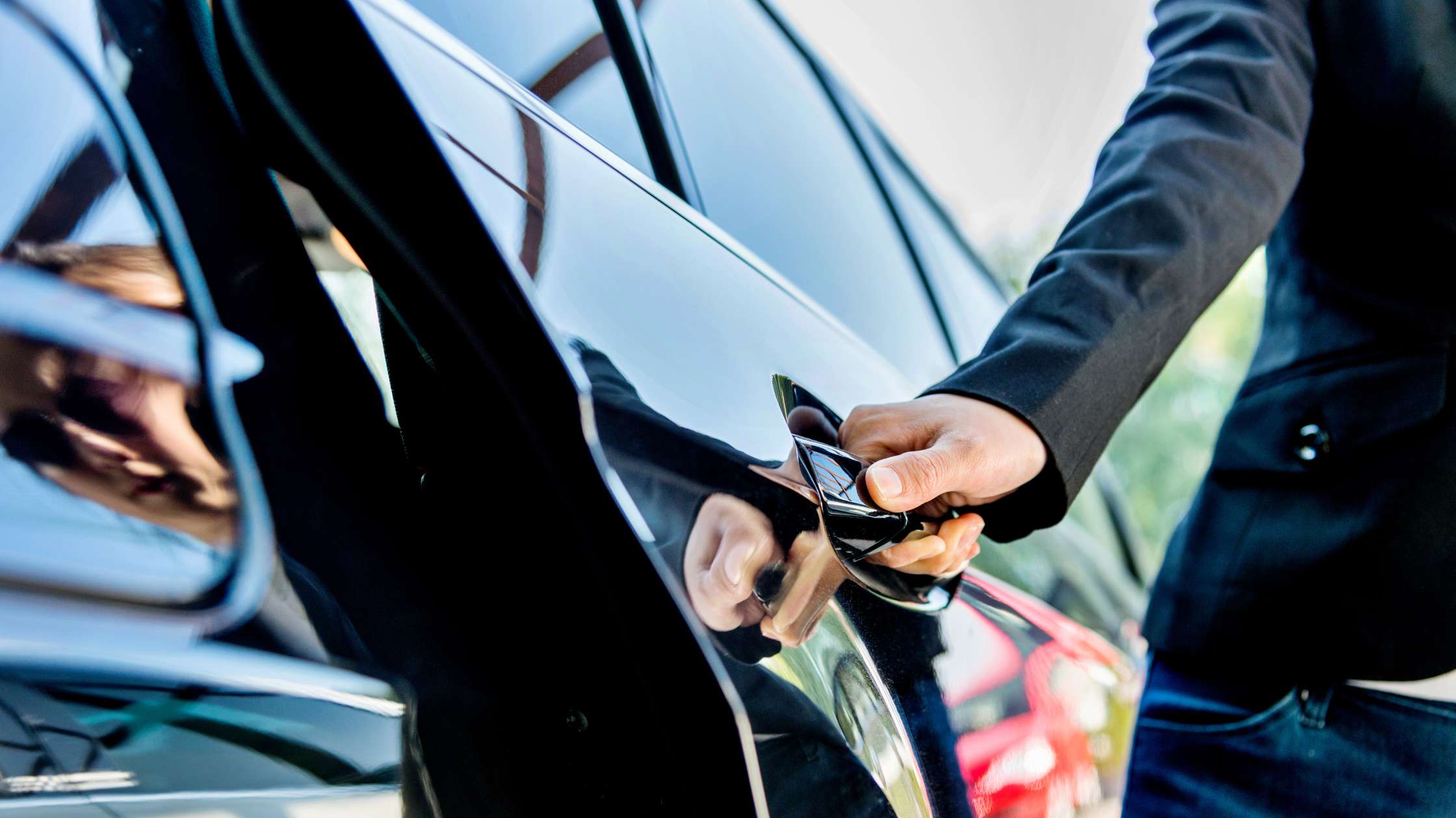 A person wearing a dark suit is opening the door of a shiny black car. The image is focused on their hand gripping the car door handle.
