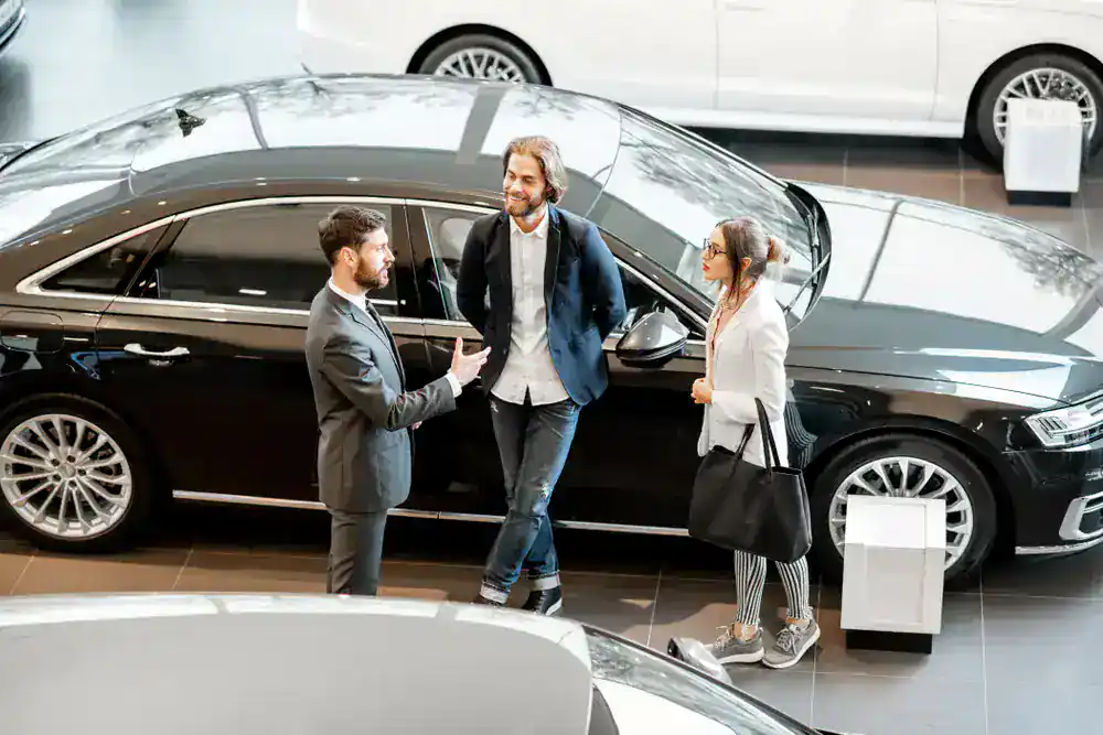 A car salesperson talks with a man and woman standing beside a black luxury car inside a modern showroom, with other cars visible in the background.