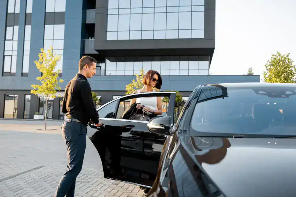 A man in black clothing opens the door of a black car for a woman wearing sunglasses and a white outfit as she steps out, with a modern glass building and trees in the background.