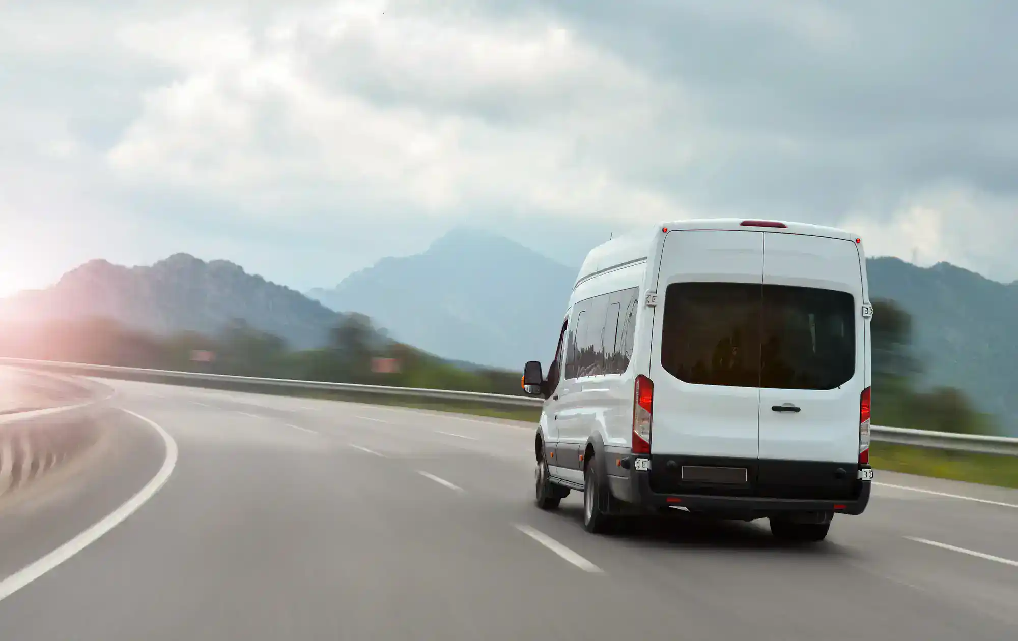 A white van drives on a curved highway surrounded by mountains and greenery under a cloudy sky, with sunlight peeking through in the distance.