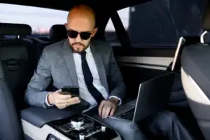 A man in a gray suit and sunglasses sits in the backseat of a car, looking at his smartphone. He has a laptop open on his lap and appears focused, suggesting he is working while traveling.